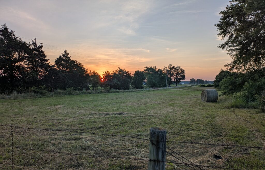 Sunrise over a rural southwestern Missouri hayfield field w/a round haybale and native trees in the background.